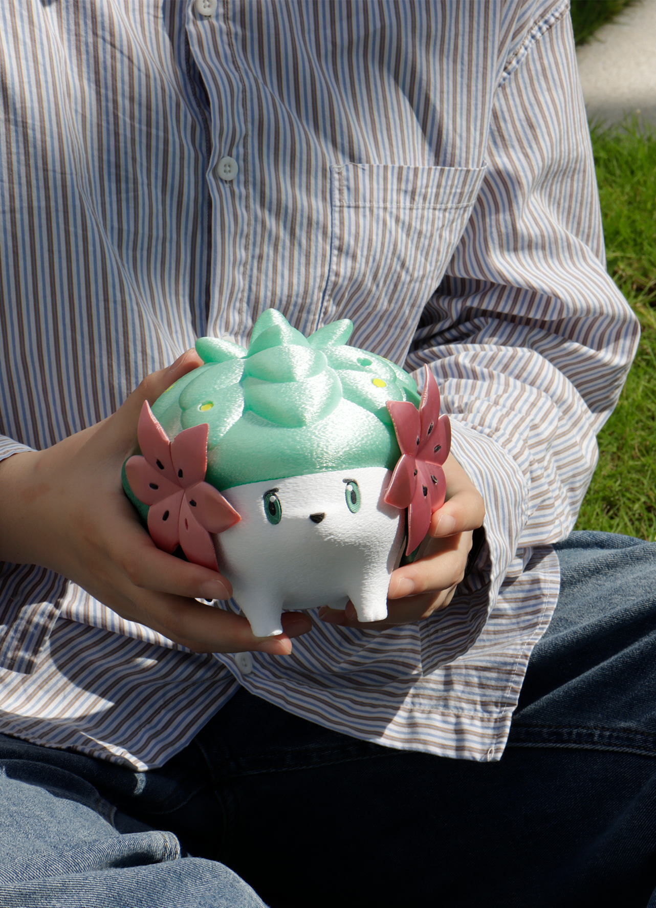 Person holding a cute 3D‑printed creature planter with mint‑green leaves and pink flowers while sitting on the grass.