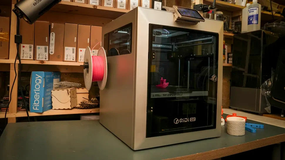 QIDI MAX4 enclosed 3D printer on a workshop workbench, with a pink filament spool mounted on its side and a small pink print inside the build chamber.